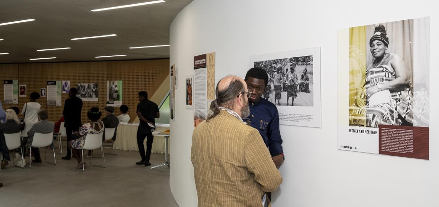 two men talk in front of posters on wall