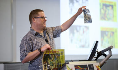 A person stood behind a podium holding up reproductions of two old hollywood posters