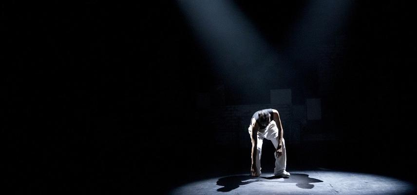 Image of performer touching one hand on the ground in spotlight on a dark stage