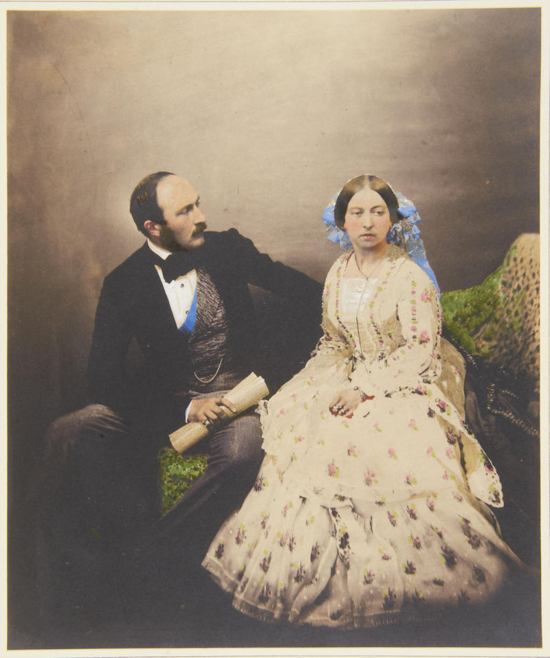 Hand-tinted photograph. Prince Albert is looking at Queen Victoria, who is looking away. The Queen is wearing a white dress and a veil.
