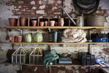 Three shelves with everyday gardening tools and similar objects, in front of a wall of rubble masonry, whose plaster has been partially peeled off.