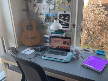 The desk of the author is facing the window. Her laptop is open. Alongside stationary, a guitar is on the desk, placed against the wall.