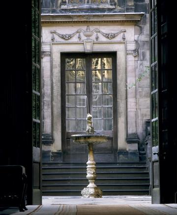 A photography taken from an interior space at Dunham Massey, looking towards a courtyard, where a fountain stands. Behind the fountain, there are the heavy glass doors of a different room that opens to the same courtyard but at a higher level (six steps).