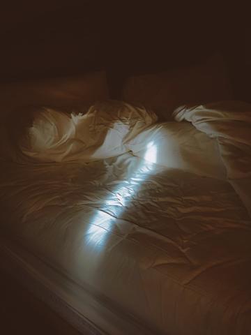 A bed with crumpled sheets. There is a small sliver of light shining onto the mattress.