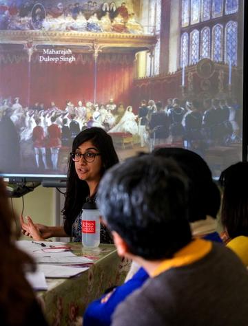 Dr Atwal has long dark hair and wears glasses. She stands in front of a powerpoint presentation depicting a painting of Queen Victoria's wedding. An arrow on the poerpoint points to Maharajah Duleep Singh who is depicted in the audience.