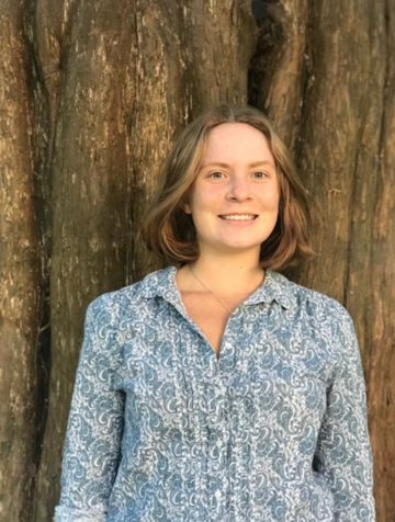 Madeleine has light brown short hair and smiles at the camera. She wears a patterned shirt and stands in front of a massive tree trunk.