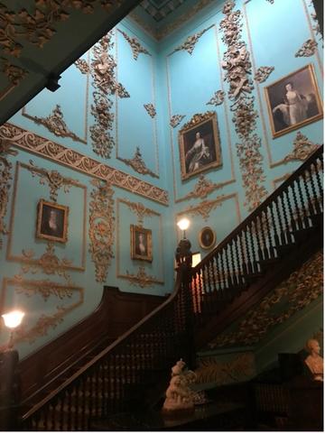 Photo from the bottom of the stairs looking up. The walls are painted in a light blue and decorated with paintings and golden mouldings. 