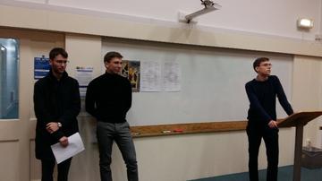 Three members of the OUBS committee giving a speech. One stands at a lectern; the other two stand by the door to the left of the photograph. The conference poster is visible in the background.