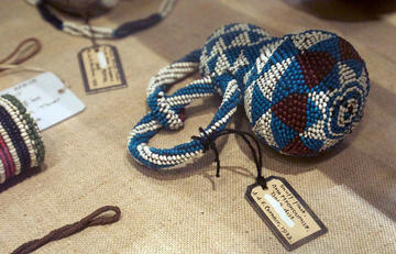 A gourd with two handles made out of blue, white and brown beads in a triangle pattern with a label hung on it lying on a cloth.
