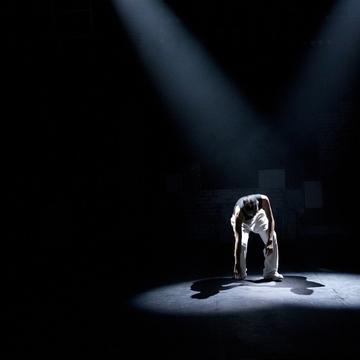 A performer under a spotlight touching one hand on the floor of a dark stage