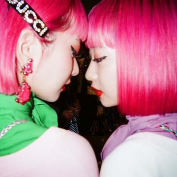 Headshot of two women with bright pink hair facing each other, close together in a romantic posture