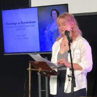 Maisie stands in front of a presentation screen and reads from a script. Maisie is blonde with long hair and wears a white shirt over a grey t-shirt and black trousers.