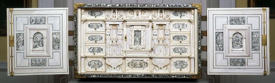 Frontal view of cabinet with doors wide open. All inner surfaces are covered with ivory inlays displaying monochrome scenes from the bible Book of Esther. The interior consists of multiple drawers and small cupboards.
