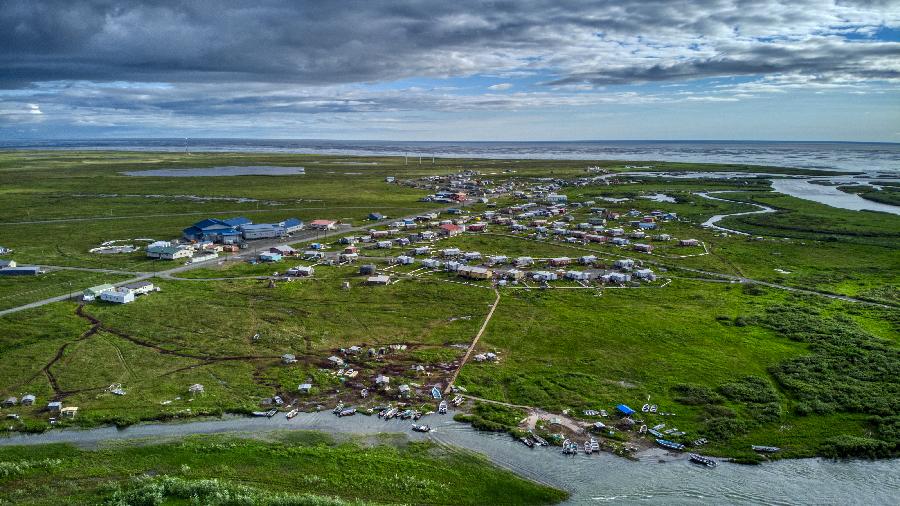 Aerial photograph of Quinhagak facing west, a Yup’ik community with dirt/gravel roads by the sea and Kanektok river, surrounded by lush green tundra in summer.